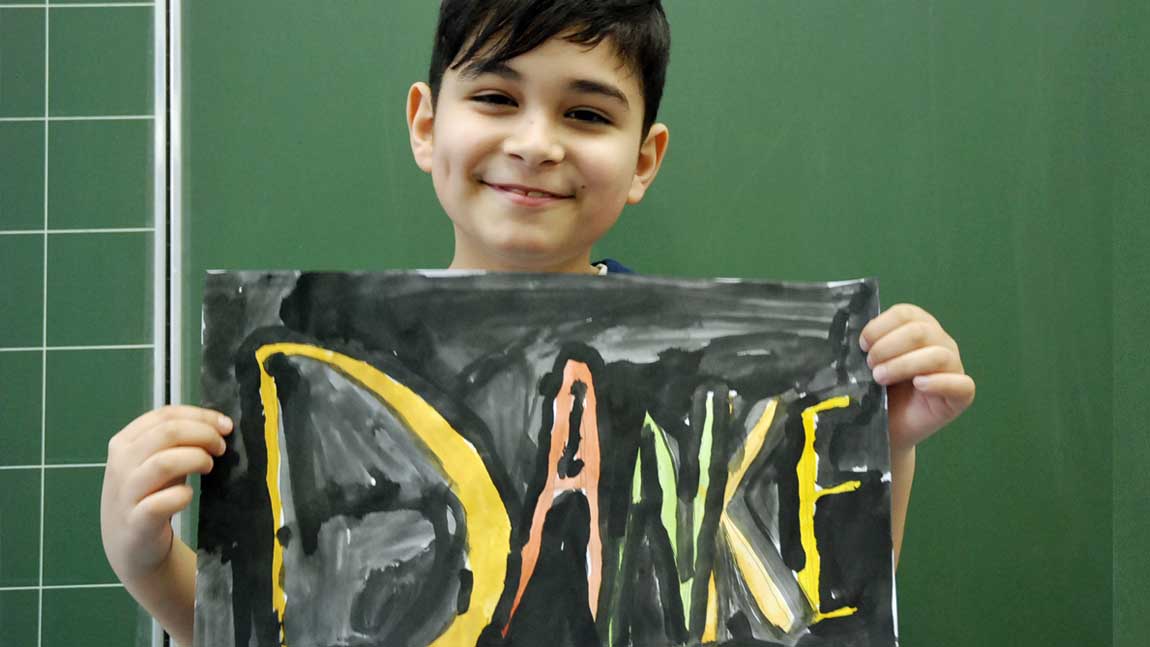 Duneklhaariger Junge steht lächelnd vor einer Schultafel und hält ein Bild vor seine Brust. Darauf steht "Danke"