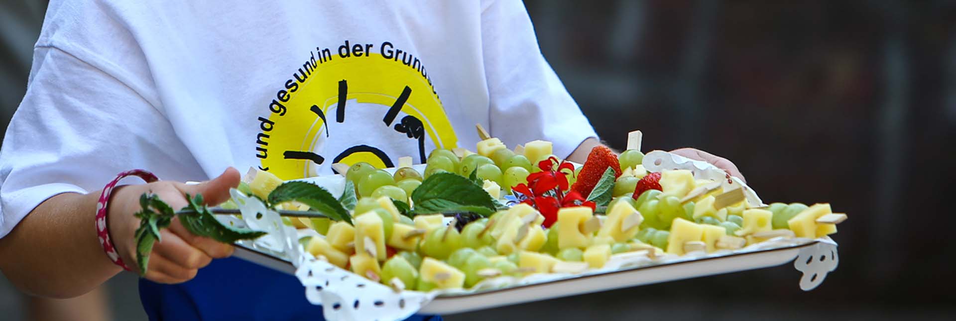 Ein Kind mit einem KLARO-Tshirt trägt ein Tablett mit Obst-Spießen