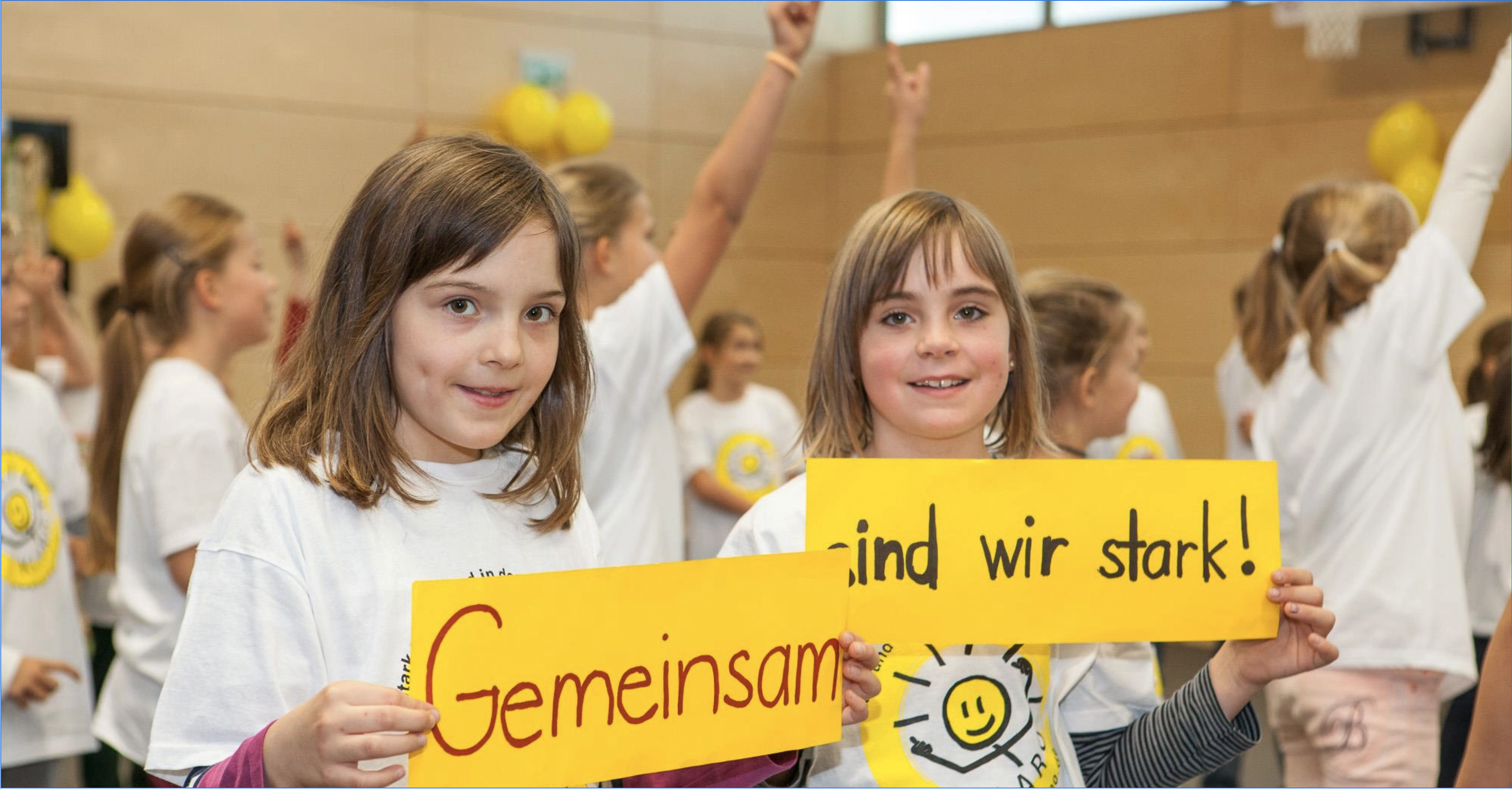 In einer Turnhalle mit Kindern halten zwei Mädchen Schilder vor sich und blicken in die Kamera. Auf den Schildern steht: Gemeinsam sind wir stark.