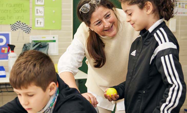 Eine brünette Frau beugt sich zu einem Mädchen in einem Klassenzimmer, sie massiert den Rücken eines Jungen