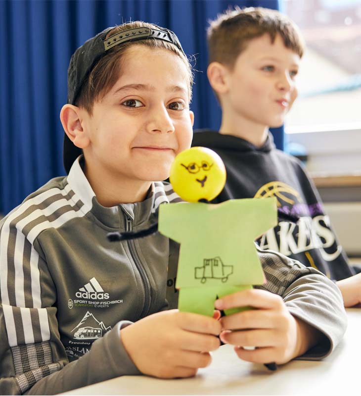 Mit KLARO gesund bleiben lächelnder Junge sitzt am Tisch im Klassenzimmer und hält KLARO in der Hand