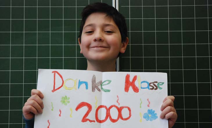 Ein 8-jähriger Junge steht frech grinsend vor einer Kamera und hält ein bunt bemaltes Plakat vor die Brust. Darauf steht: Danke Klasse2000