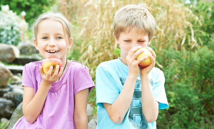 Ein Junge und ein Mädchen im Grundschulalter sitzen in einem Garten nebeneinander und essen genüsslich Äpfel.