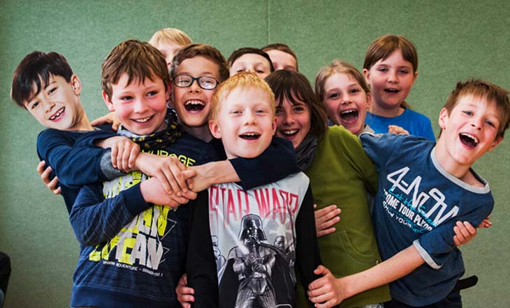 So gelingt Teamarbeit Eine Gruppe von elf lachenden Kindern im Grundschulalter steht in einer Turnhalle sehr eng zusammen. Die Kinder halten sich gegenseitig fest.
