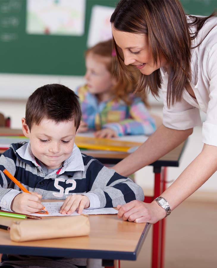 Gesundheitsbildung die Spaß macht Eine junge brünette Lehrerin beugt sich zu einem sitzenden, jungen Schüler. Der Junge grinst verschmitzt und schreibt in sein vor sich liegendes Heft.