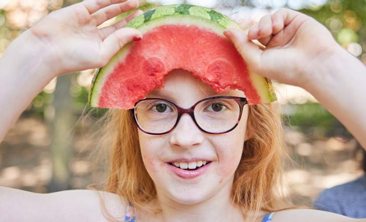 Melone erfrischt und schmeckt Ein frech grinsendes, rothaariges Mädchen mit Brille steht im sommerlichen Pausenhof und hält sich ein Melonenstück an die Stirn.