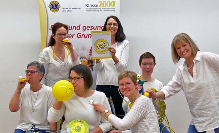 Top vorbereitet mit unseren Schulungen Eine gruppe von sieben fröhlichen Frauen in weißen Pullovern experimentiert mit Material aus dem Klasse2000-Programm: Luftballons, Stethoskop, Atemtrainer.