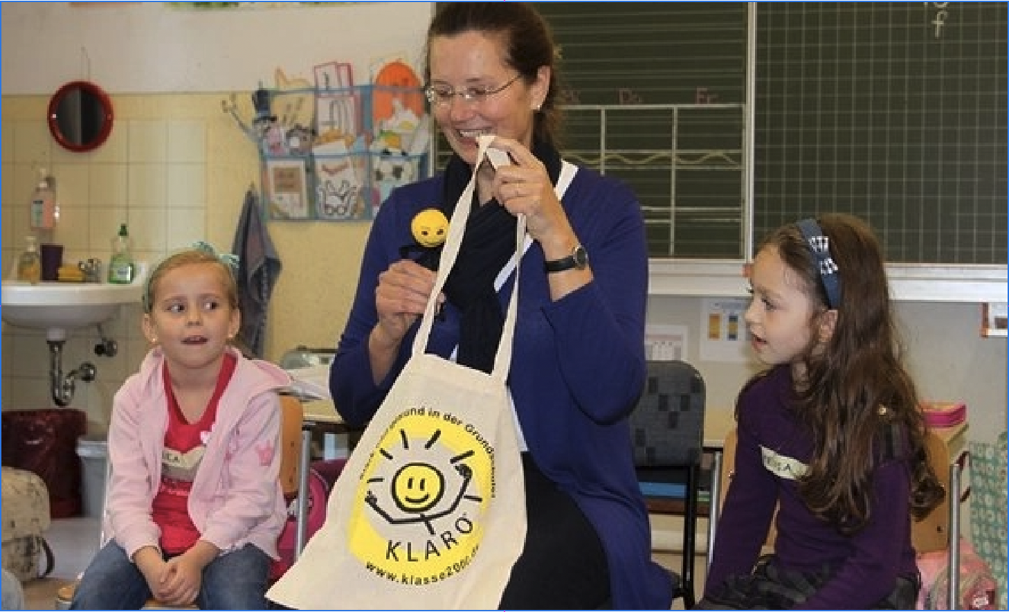 KLARO stellt sich vor Eine dunkelhaarige, mit Brille Frau sitzt in einem Klassenzimmer auf einem Stuhl zwischen zwei Mädchen. Die beiden Mädchen blicken gespannt auf die Frau, die KLARO aus ihrer Stofftasche holt.