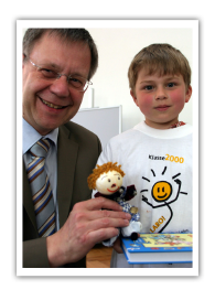 Karl Freller mit 150.000stem KLARO Kind Ein Mann in Anzug hält lächelnd eine KLARO Puppe in der Hand. Neben ihm steht ein Junge mit weißem KLARO T-Shirt