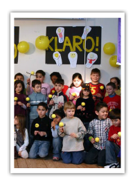 Lions fördern Klasse2000 seit Anfang an Eine Gruppe von Kinder hält gebastelte KLAROs in der Hand. Hinter ihnen an der Wand sind gelbe Ballons und ein Schild mit der Aufschrift KLARO.