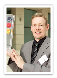 Thomas Duprée erhält Auszeichnung Ausgewählter Ort 2012: Im Wettbewerb „365 Orte im Land der Ideen“ Der Geschäftsführer von Klasse2000 trägt einen grauen Anzug und hält stolz die Auszeichnung "365 Orte im Land der Ideen"