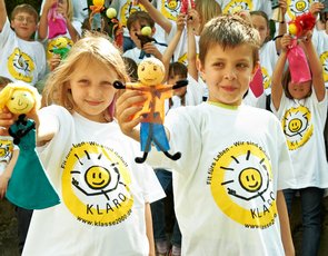Ein Mädchen und ein Junge in KLARO-T-Shirts halten gebastelte KLAROs in die Kamera. Dahinter sind weitere Kinder aus ihrer Schulklasse zu sehen.