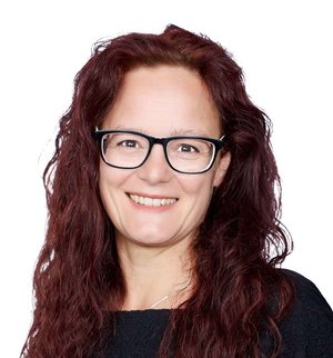 Kerstin Waider Porträt vor weißem Hintergrund, von einer Frau mit langen lockigen roten Haaren, Brille und schwarzem T-Shirt, die in die Kamera lächelt.
