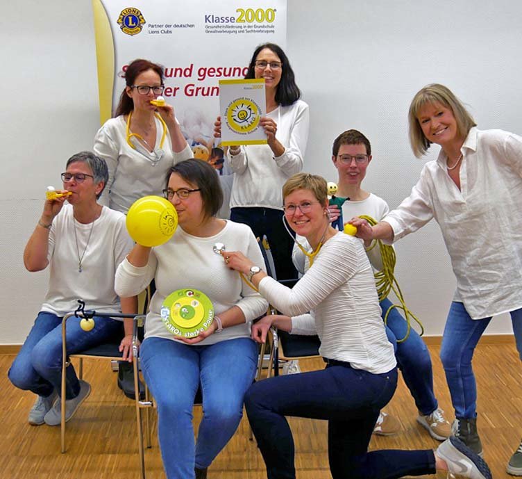 Eine Gruppe von sieben fröhlichen Frauen in weißen Pullovern experimentiert mit Material aus dem Klasse2000-Programm: Luftballons, Stethoskop, Atemtrainer.