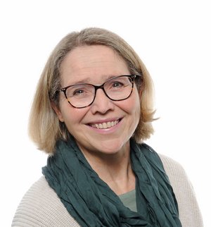 Annedore Fricke Porträt vor weißem Hintergrund, von einer Frau mit blonden, schulterlangen Haaren, Brille, beigem Strickpullover und petrolfarbenem Schal, die über ihre rechte Schulter in die Kamera lächelt.