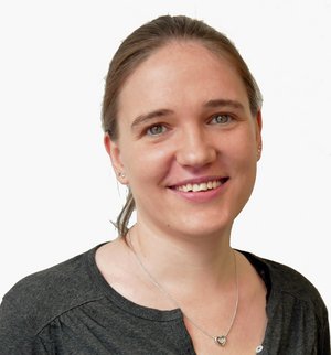Julia Feldmann Porträt einer jungen Frau mit Pferdeschwanz und mit grauem Longsleeve und Halskette, die in die Kamera lächelt.