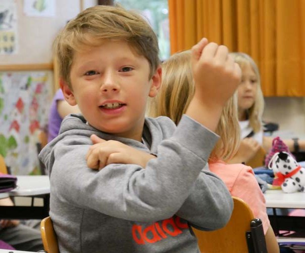 Stark und gesund aufwachsen Ein blonder Junge im Grundschulalter sitzt im Klassenzimmer, lächelt in die kamera und fühlt seinen angespannten Bizeps.