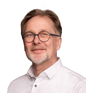 Dirk Höcherl Porträt vor weißem Hintergrund, von einem Mann mit kurzen grauen Haaren, Bart und Brille, der ein weißes Hemd trägt und in die Kamer lächelt.