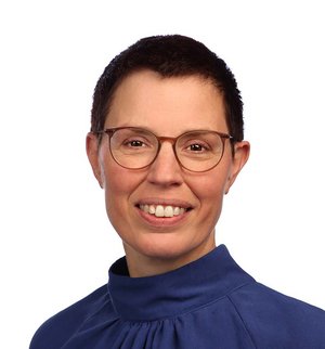 Petra Suer-Meiners Porträt vor weißem Hintergrund, von einer Frau mit kurzen braunen Haaren, Brille und blauer Bluse, die über ihre linke Schulter in die Kamera lächelt.
