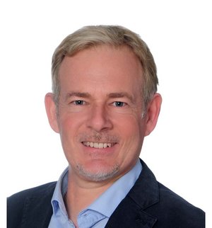 Porträt vor weißem Hintergrund, von einem Mann mit blonden kurzen Haaren und Kinnbart, der ein hellblaues Hemd und ein dunkelblaues Sakko trägt und frontal in die Kamera lächelt. 